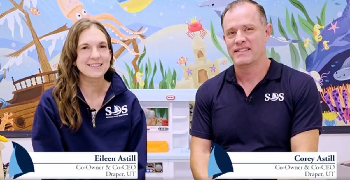 Corey and Eileen Astill, co-owners and co-CEOs of Success On The Spectrum in Draper, Utah, seated in front of a colorful underwater-themed mural.